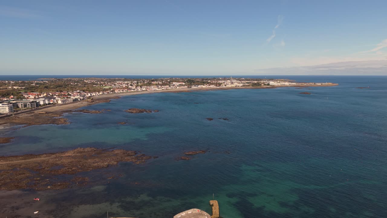 High drone footage over Belle Greve Bay Guernsey at high tide flying out over Salerie Corner and showing rocks,beach ,clear blue water and distant views of St Sampson on sunny day