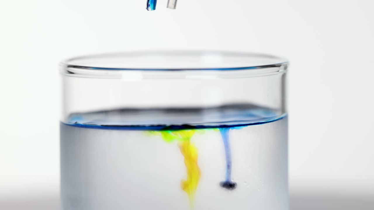 Pipettes release colored drops into clear water, creating swirling diffusion patterns under bright studio lighting