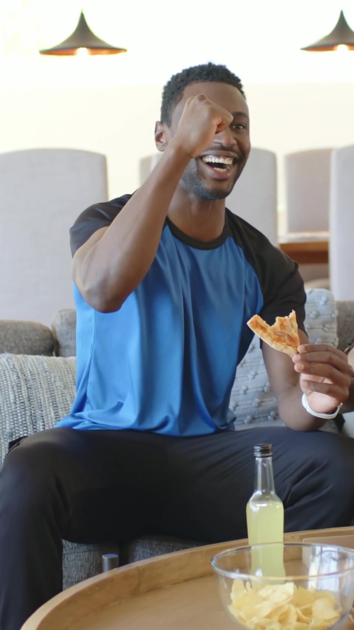 Video verticale: Uomo che esulta, con una maglietta raglan blu, sul divano, che reagisce a un gol in TV e tiene in mano una pizza.