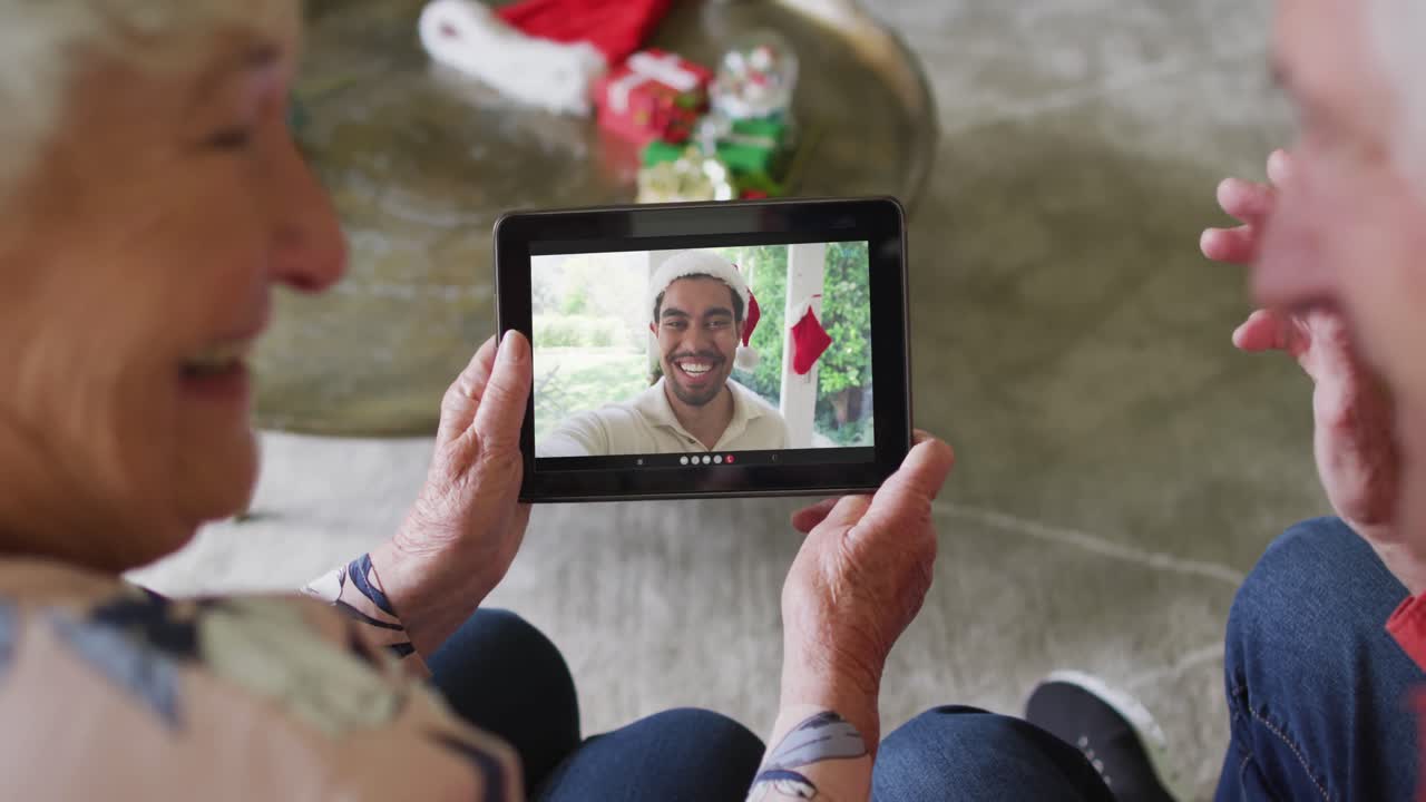 pareja de ancianos caucásicos sonrientes usando una tableta para una videollamada de navidad con un hombre en la pantalla