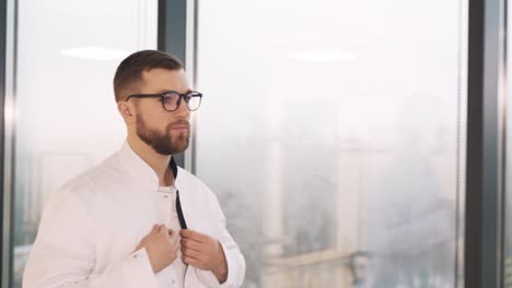 Portrait-of-a-male-doctor-walking-past-panoramic-windows-in-a-modern-clinic-and-wearing-a-white-coat