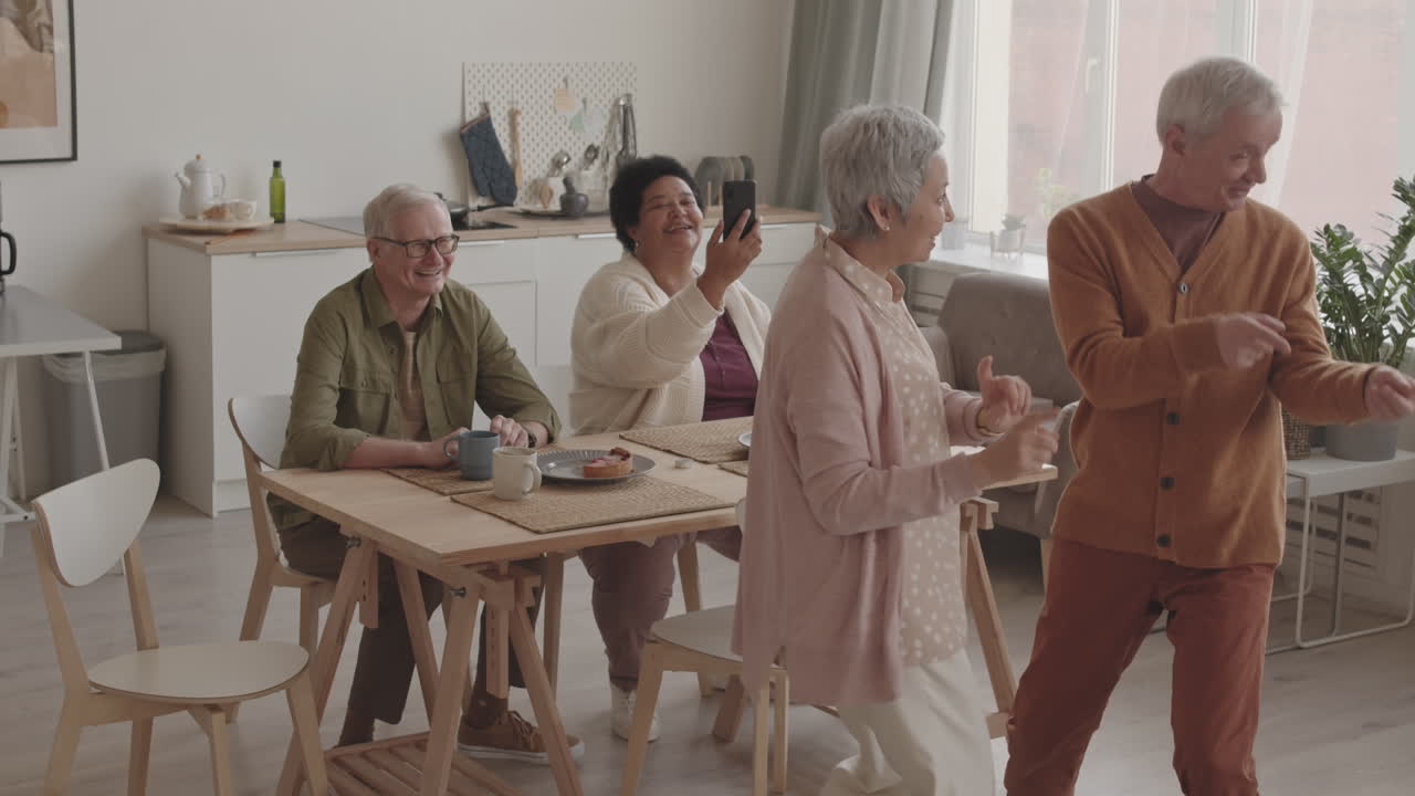 High angle medium long of grey-haired Asian woman dancing with her senior Caucasian husband, their diverse friends filming them on smartphone sitting at dinner table