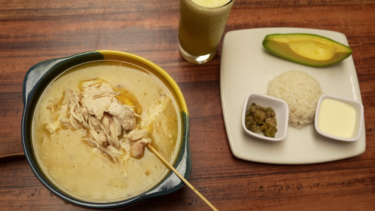 Overhead view of creamy ajiaco soup paired with rice, avocado, and flavorful sides on a wooden table. Warm lighting enhances this enticing Colombian dish presentation