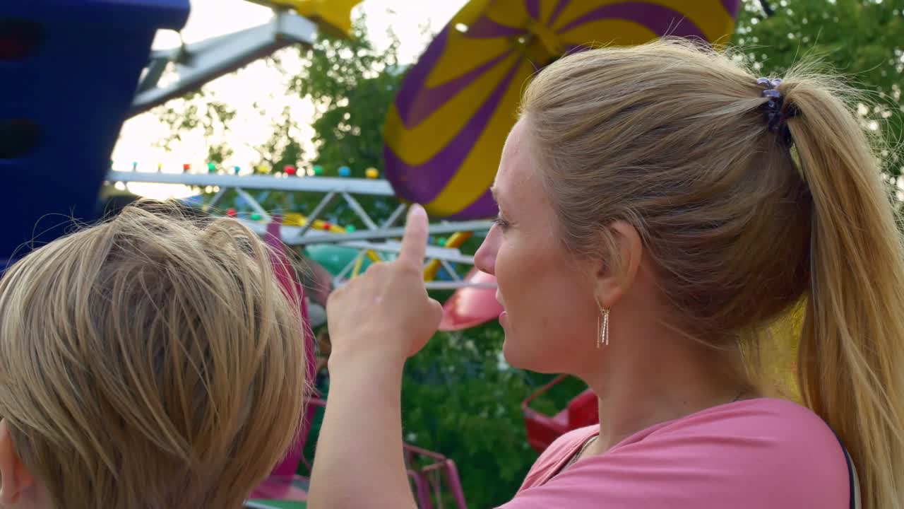 madre e hijo miran un carrusel rápido girar en un parque de atracciones, de pie en la fila discutiendo un paseo divertido. madre e hijo están teniendo un gran tiempo en la feria