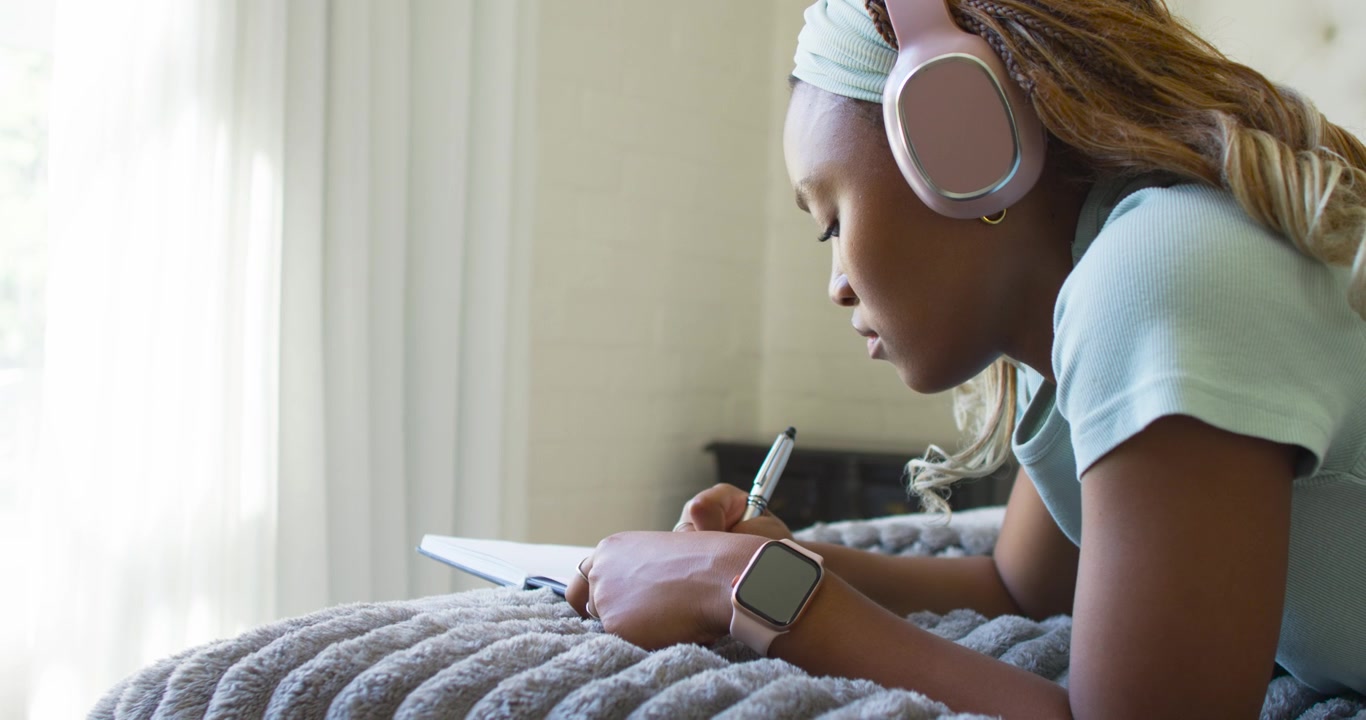 African female student lying on bed by window, listening on headphones and taking notes in notebook