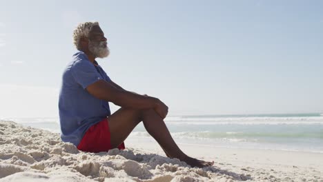 hombre afroamericano mayor sentado y mirando lejos en una playa soleada