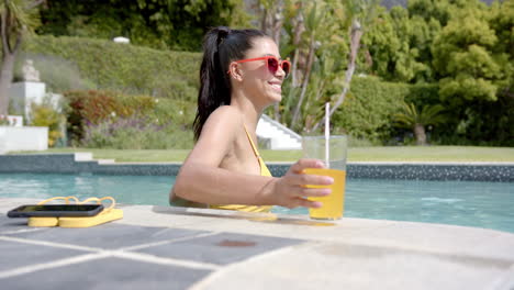 feliz adolescente biracial con gafas de sol de pie en la piscina soleada con bebida, cámara lenta