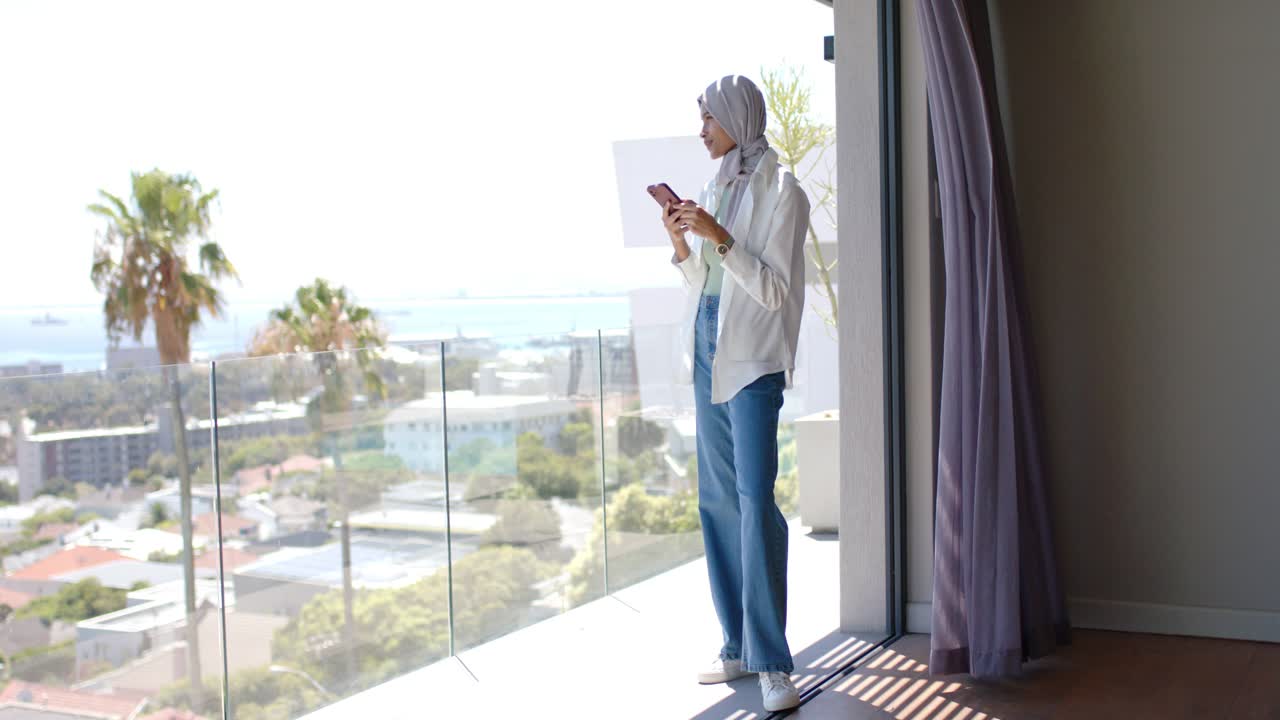 Donna africana con hijab in piedi sul balcone che tiene in mano telefono e un foglio, controllando e guardando l'oceano, con spazio per il testo.
