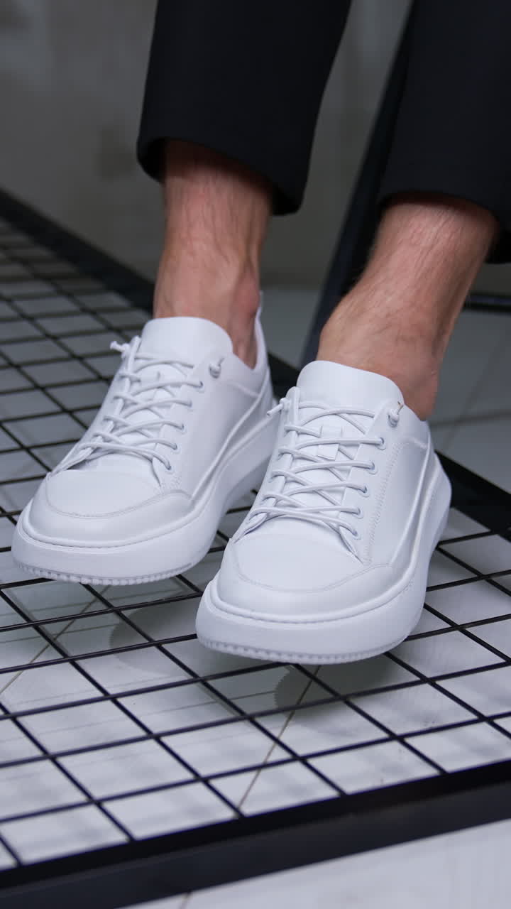 Modern convenient white sport shoes. Male model sits on chair keeping his feet in stylish sneakers close to camera. Vertical video