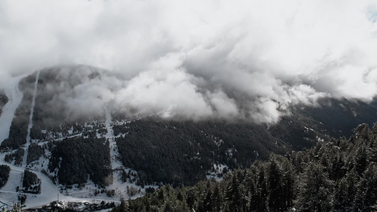 Aerial panorama between trees of a valley in cloudy alpine snowy mountain with clouds