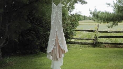 elegant designer wedding dress hanging from a pine tree branch in the yard of a acreage with a wood fence in the back ground at the strathmere wedding centre and spa