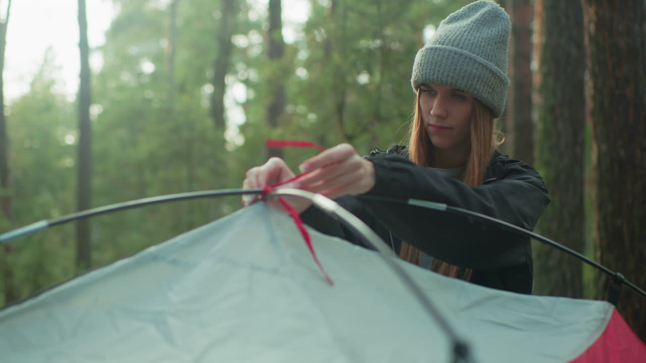 Girl wearing beanie ties red rope on flexible tent pole while smiling in forest as sunlight filters through trees during morning camping setup in wilderness with soft focus on background foliage