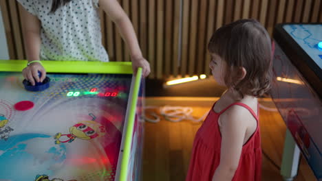 Niña-Pequeña-Viendo-A-Los-Niños-Jugando-Hockey-De-Aire-En-La-Sala-De-Juegos-De-Arcade,-Niños-Golpeando-El-Disco-De-Plástico-Con-El-Delantero-En-Cámara-Lenta