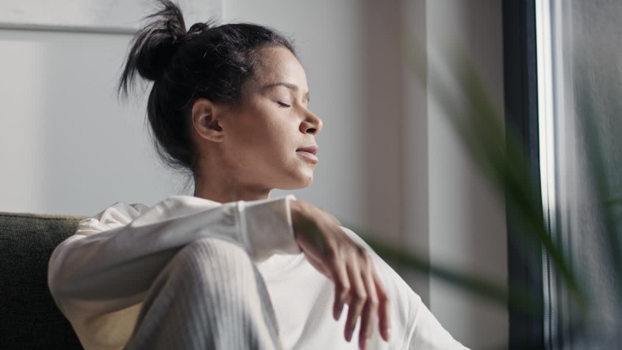 자연스러운 여성은 집에 앉아서 눈을 감고 해를 즐기고 있습니다.