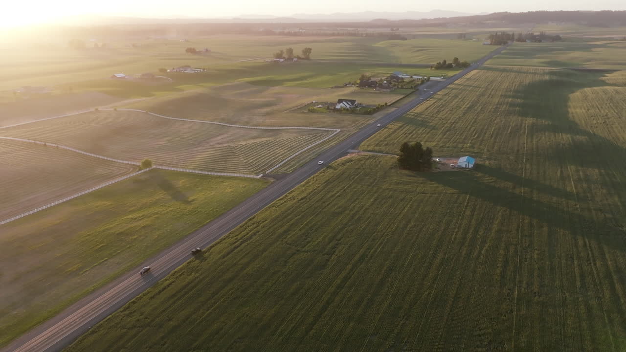 Cars drive down a country road surrounded by big open fields. The sun is low, casting shadows across the land