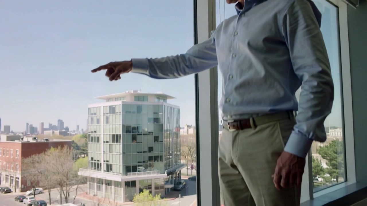 Homme d'âge moyen en chemise bleue pointant du doigt, apercevant un bâtiment en verre par la fenêtre du bureau, avec espace de copie.