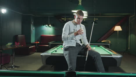 young man in white shirt and grey trousers sits on green pool table holding cue stick and sipping lemon drink. pool hall setting with stylish overhead lighting and scattered billiard balls