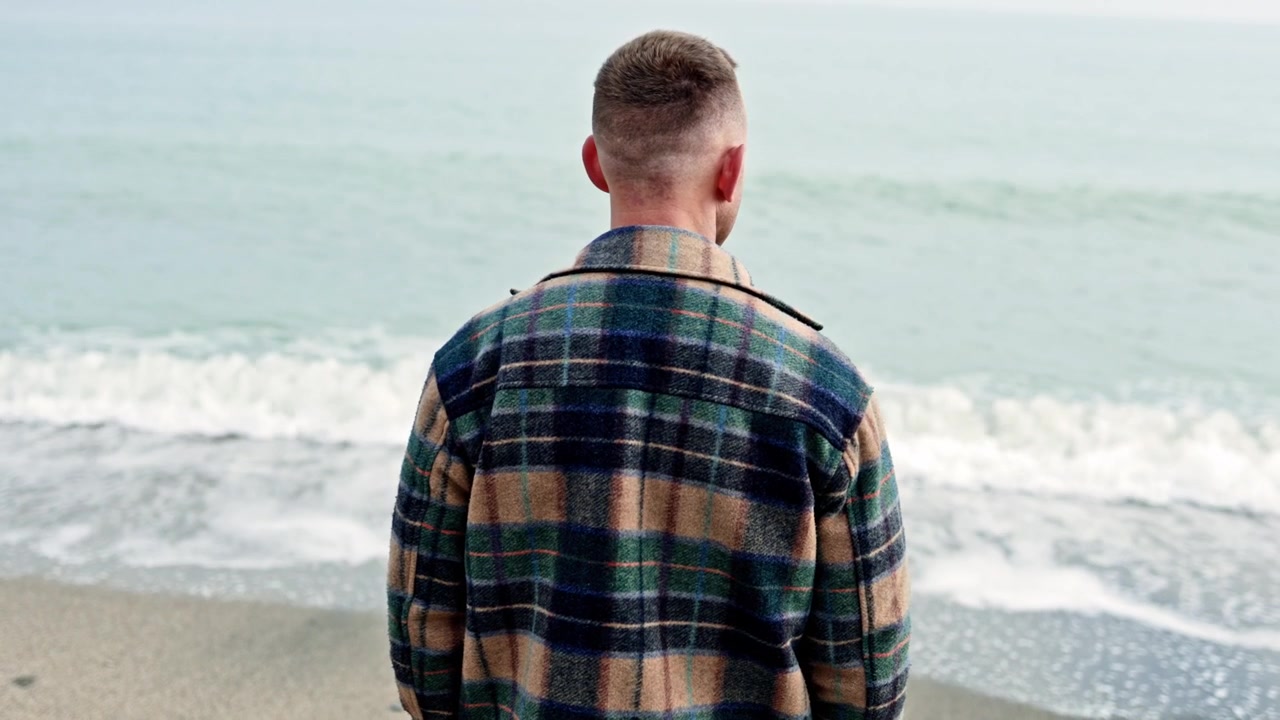 A young man stands on the shore and looks into the sea. A man walks by the sea in autumn. A man in a coat stands and looks into the sea.