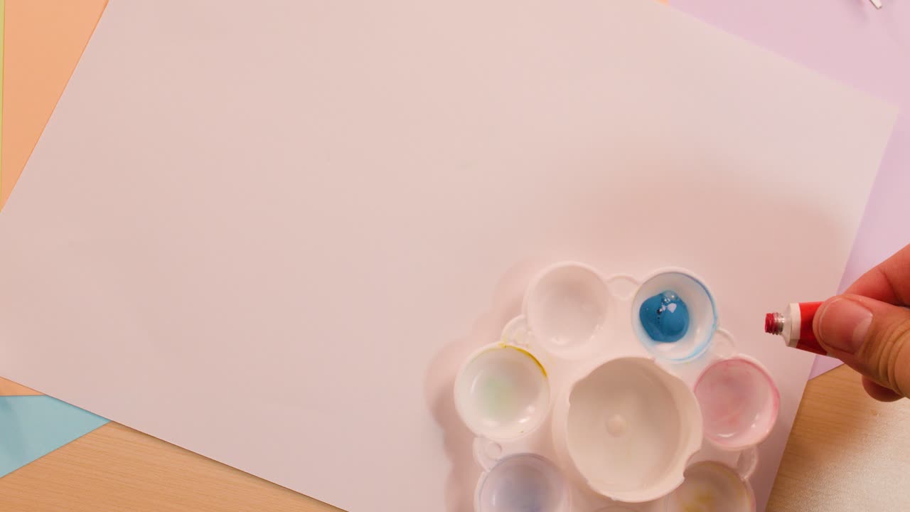 Hands squeeze blue, red, and yellow paint onto palette beside blank paper under soft lighting