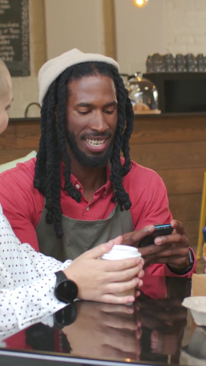 Vertical video: Approaching barista in apron leaning over table at cafe, tapping phone to show them