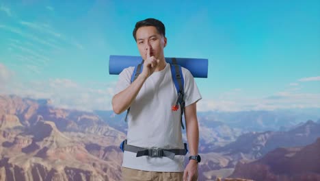 asian male hiker with mountaineering backpack looking at camera and making shh gesture while traveling at the top of mountain