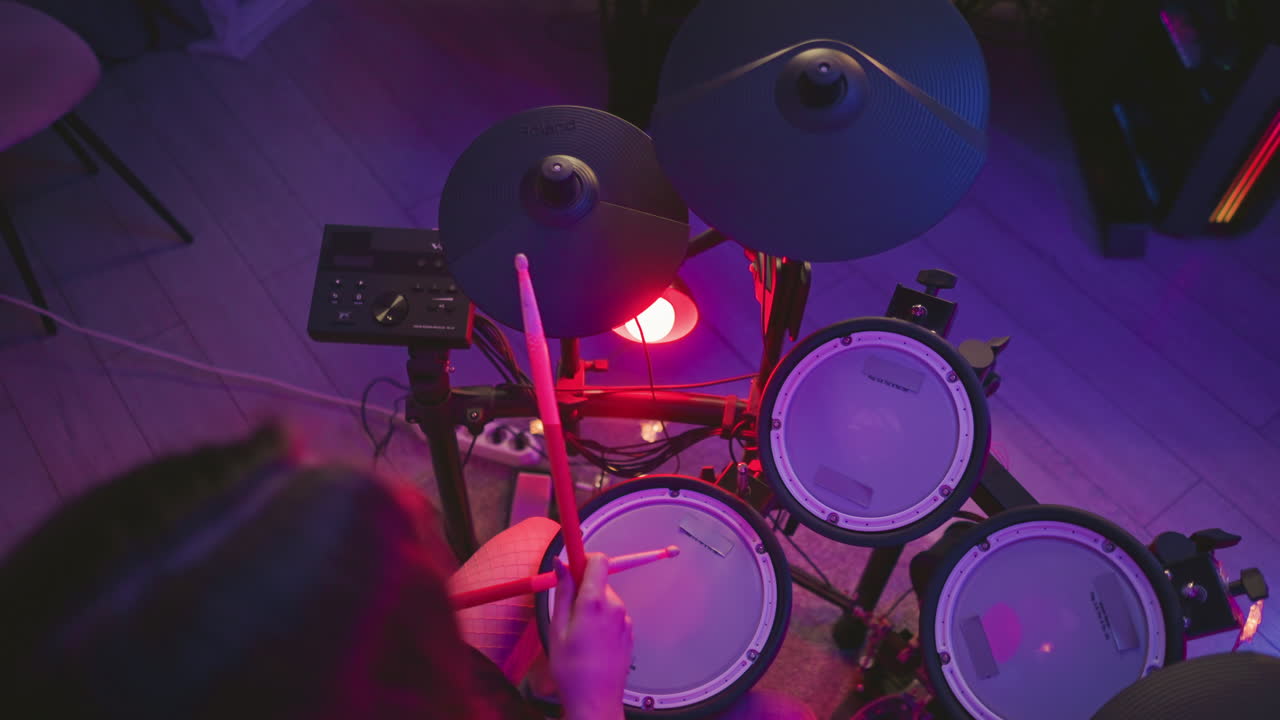 Overhead view of woman playing electronic drum kit in dimly lit room with colorful purple and red lighting, holding drumsticks and striking pads near control module and floor cables