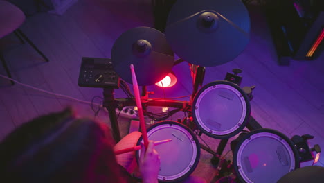overhead view of woman playing electronic drum kit in dimly lit room with colorful purple and red lighting, holding drumsticks and striking pads near control module and floor cables