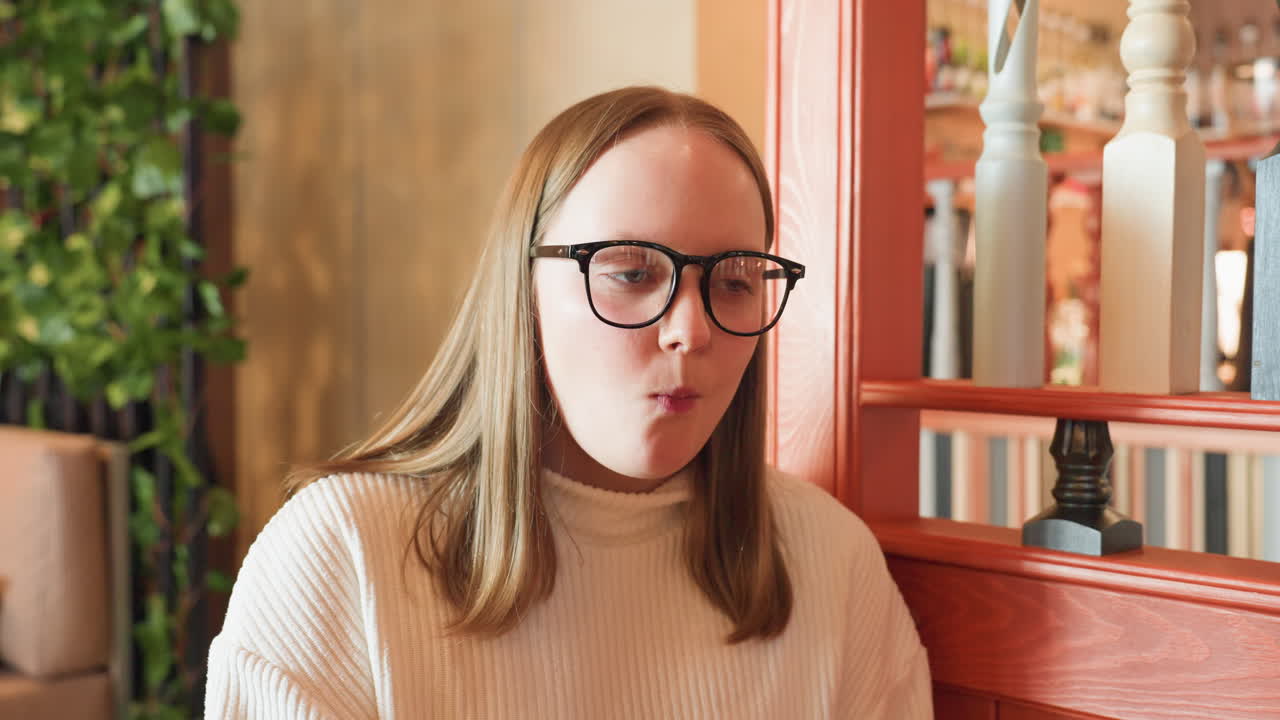 young woman in glasses enjoys sweet dessert with content expression in cozy cafe setting, captured mid bite with hand near mouth, warm tones and blurred background