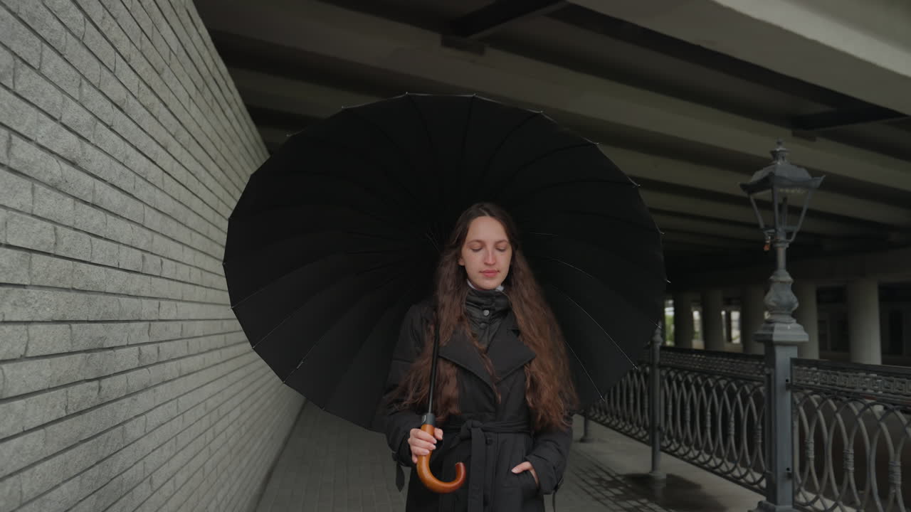 Donna con ombrello nero che cammina in un sottopassaggio, ritratto urbano malinconico con lunghi capelli ricci e cappotto nero, pendolare pensierosa ferma accanto a una ringhiera di metallo, cielo nuvoloso e marciapiede bagnato dalla pioggia.