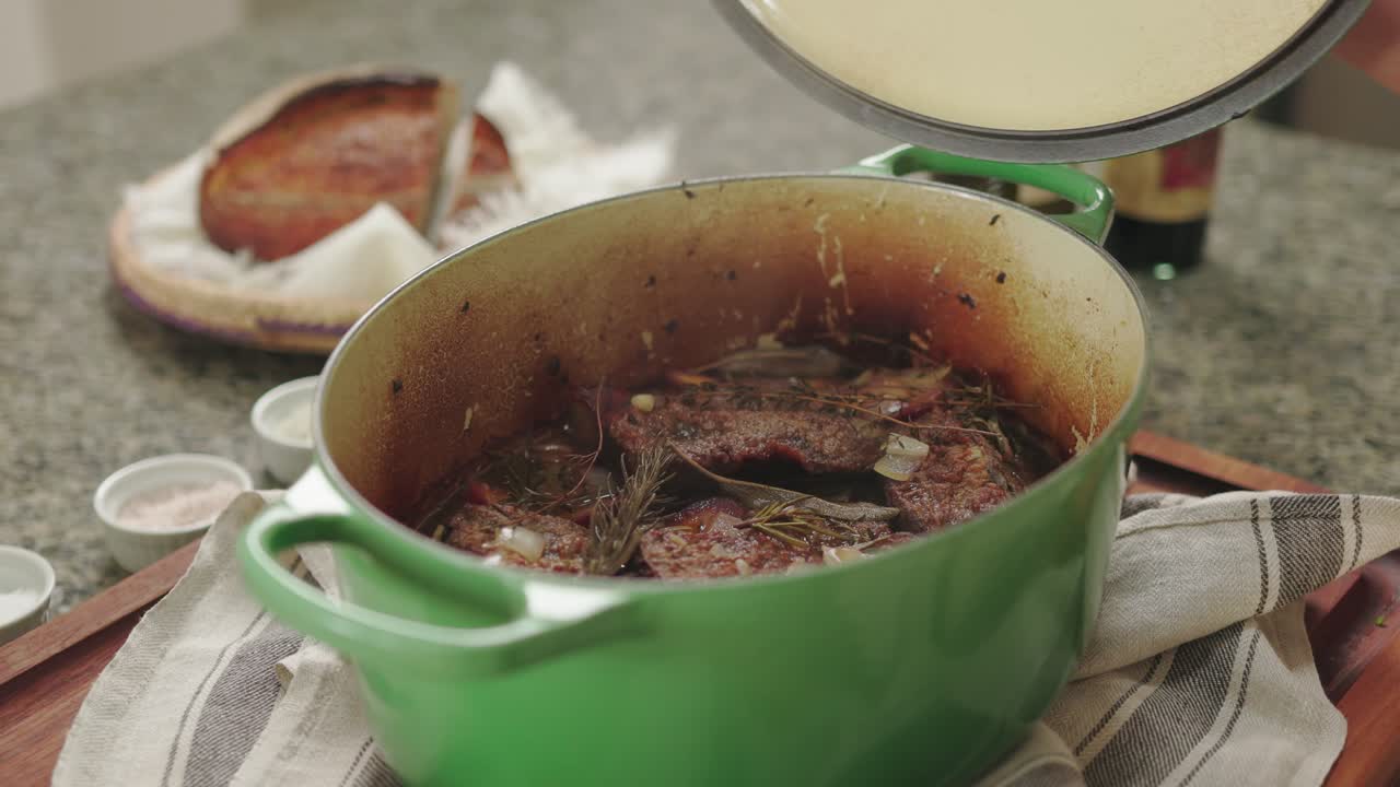 una olla de olla verde humeante con una tapa abierta, asado tierno, hierbas y pan en la parte de atrás