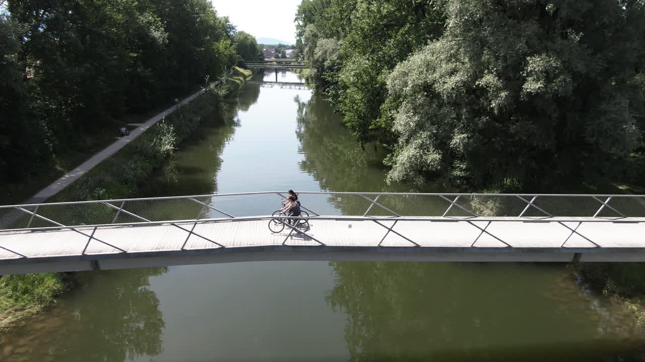 Aerial shot of two women biking over a bridge at a riverwith a lot of green trees
