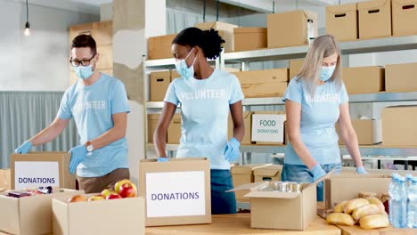 multiethnic group of volunteers in facial mask packing boxes with food and clothes in charity warehouse