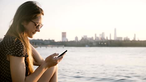 chica caucásica pensativa con gafas de sol usando una aplicación de redes sociales de teléfono inteligente, disfrutando de una increíble puesta de sol en la playa de la ciudad 4k