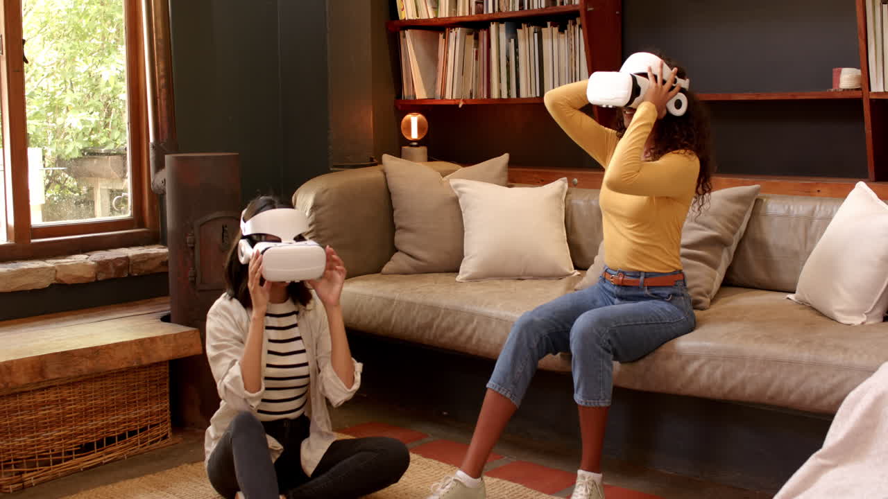 Multiracial female friends enjoying virtual reality experience at home, sitting on couch and floor