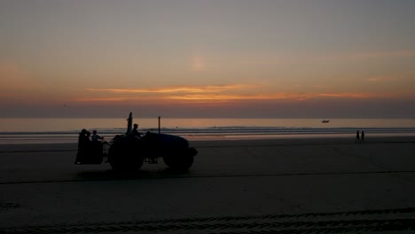 tractor passing on the beach at sunset with people in the distance, seagulls, a fishing boat in the distance and surfers in the water