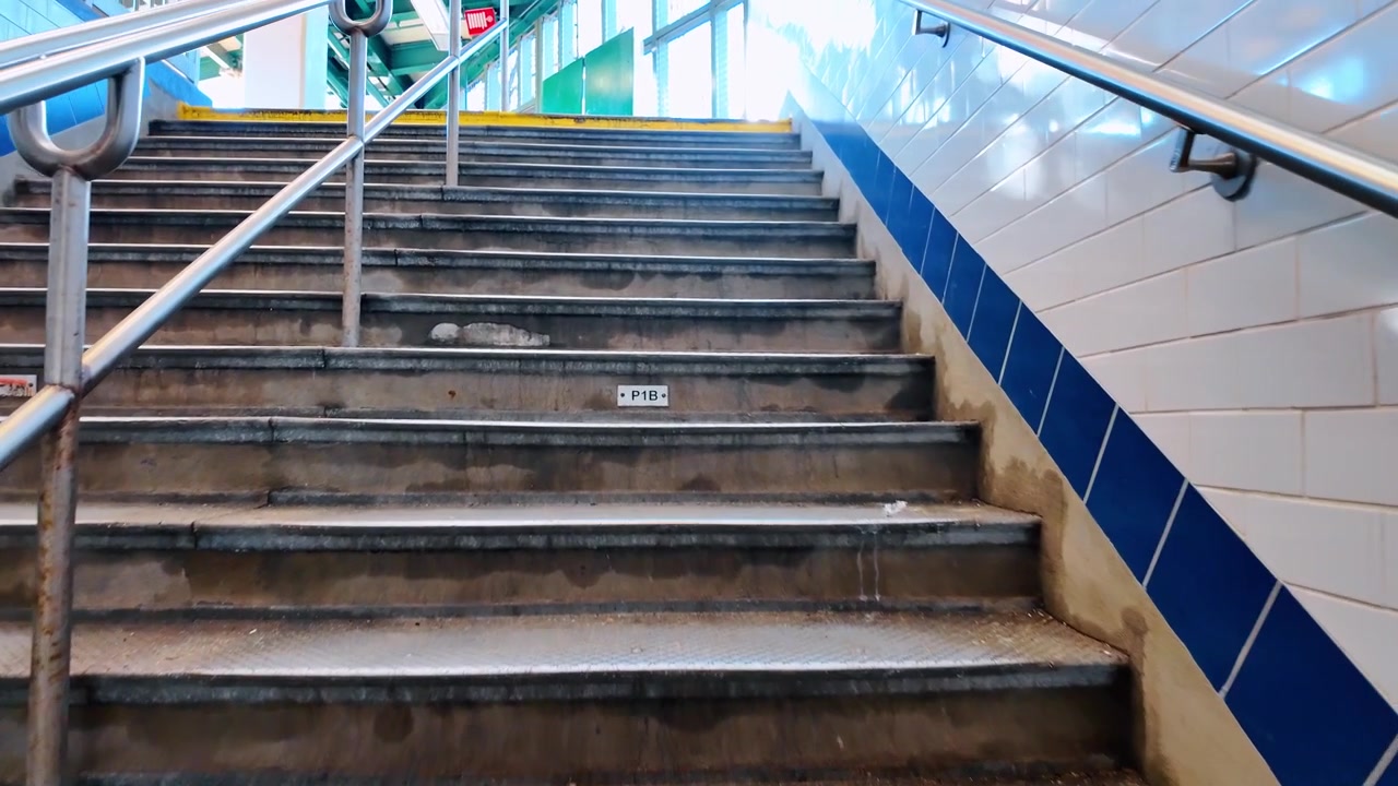 U-Bahn-Treppe mit Blick nach oben, weißen Fliesenwänden und Mosaikkunst. Eine Untersicht zeigt eine leere Treppe mit Metallgeländern, die zu einer Bahnsteigplattform führt.