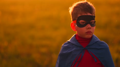 the boy in the mask and cape of a super hero at sunset in a field.