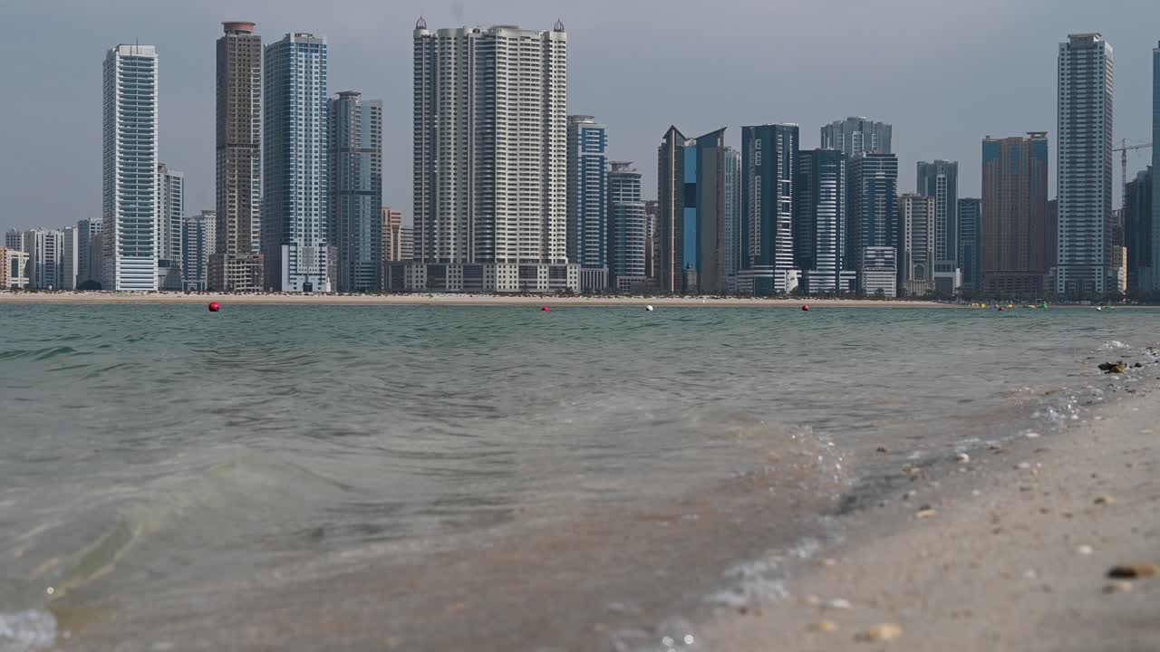 sharjah: agua de mar golpeando y rompiendo en una playa, olas de mar y rascacielos urbanos de la ciudad de sharjah en el fondo