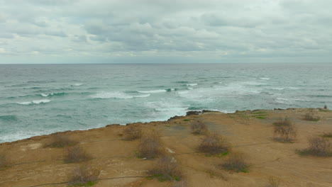 Aéreo:-Escena-Tranquila-A-Lo-Largo-De-La-Costa-De-Chipre,-Donde-Las-Suaves-Olas-Del-Mar-Mediterráneo-Bañan-Una-Costa-Escarpada-Salpicada-De-Escasa-Vegetación-Bajo-Un-Cielo-Nublado