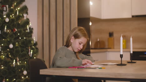 girl in green sweater folding a letter and inserting it in a envelope sitting at a table in a room decorated with a christmas tree, then gets up from the chair and leaves the room