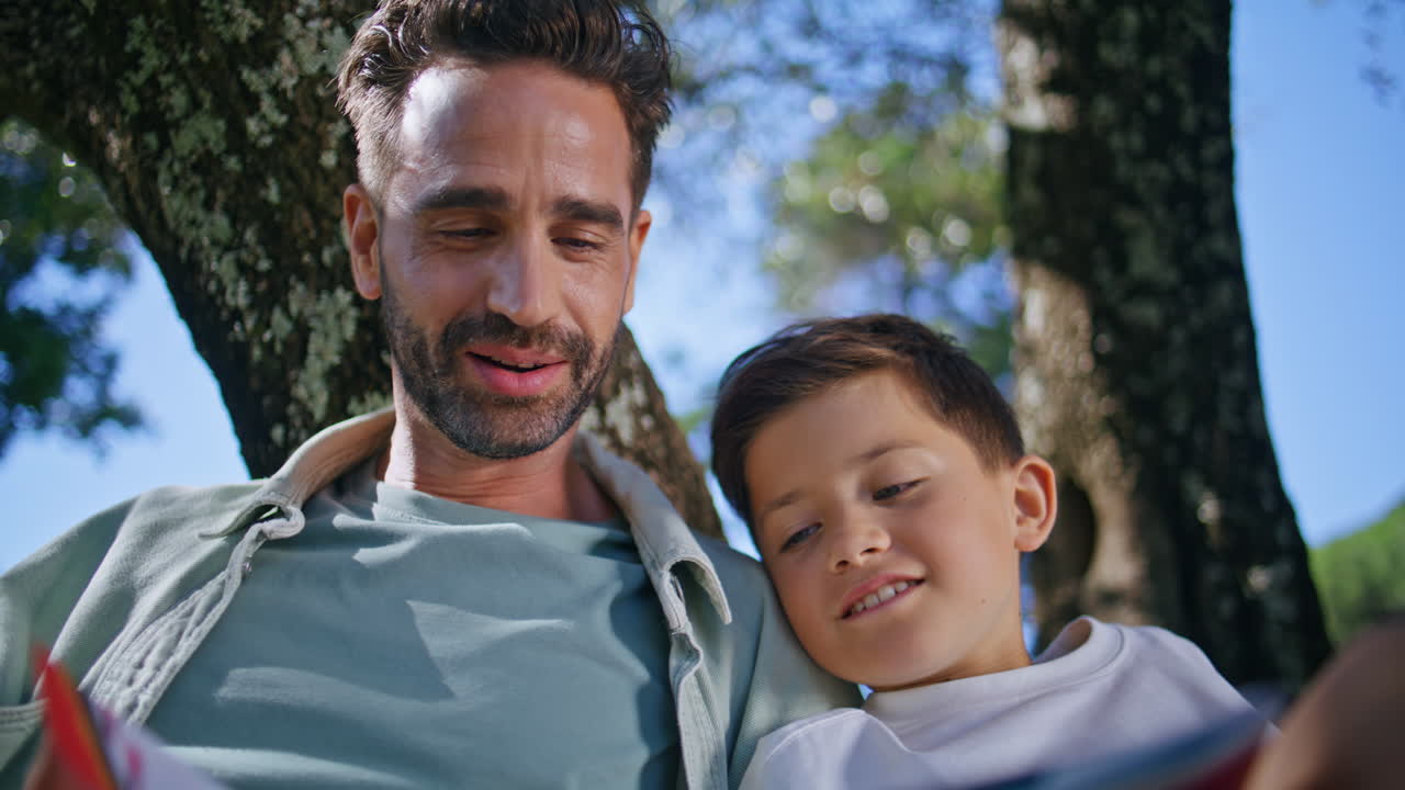 Father son flipping pages sitting under tree park closeup. Happy family reading