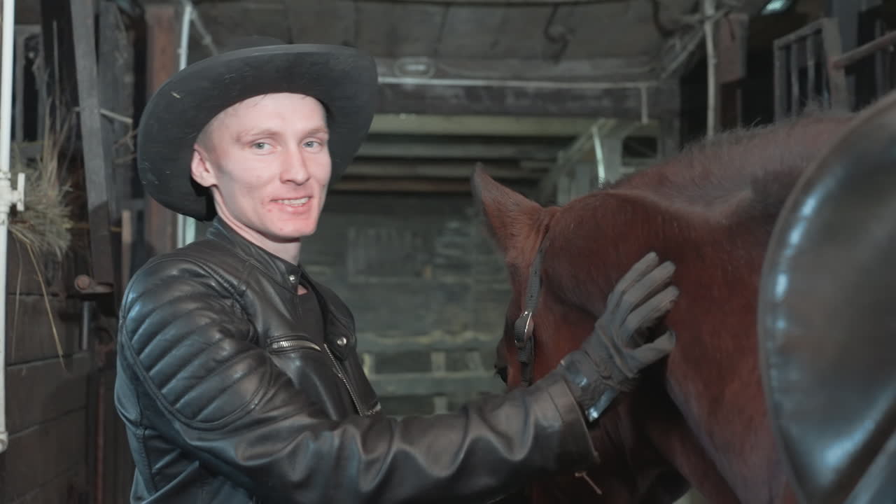 Hvid cowboy sadler hest i stald, klargør udstyr med læderjakke og handsker, rolig berøring og opmuntrende smil, staldbaggrund og rustik sadelforberedelse, partnerskab før ridetur.