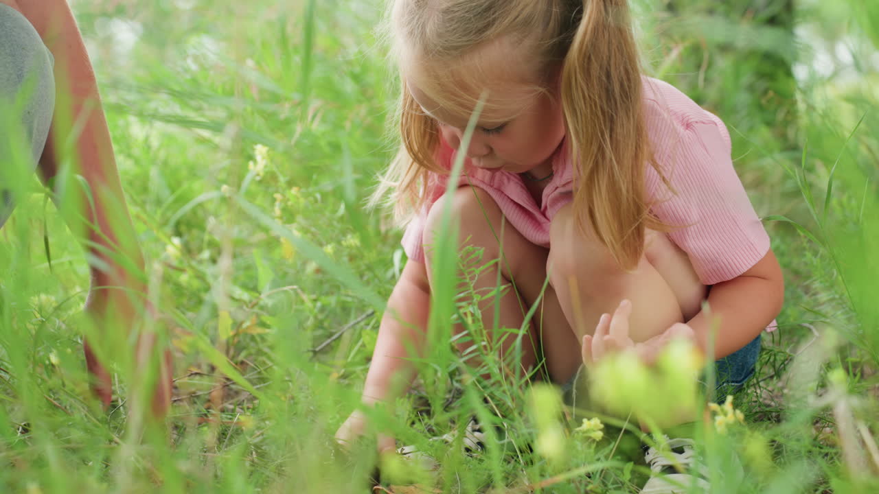 Una chica blanca agachada en la hierba explorando pequeñas plantas y flores, luz de verano suave, una mano adulta guiándola, primer plano de las manos y los tallos, ambiente tranquilo de prado en el patio trasero, juego slow living y sensorial