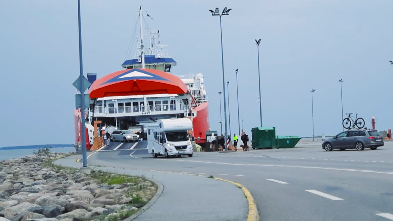 Cars and vehicles driving off a ferry that has docked called Piret in Virtsu Estonia Harbor during summertime in slow motion while the sky is cloudy. Workers guiding the cars are guiding the vehicles.