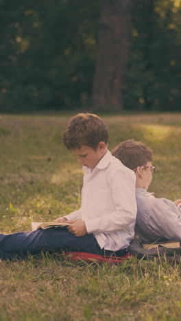 cute school friends learn textbooks sitting back to back on fresh grass lawn after lessons in sunny spring park