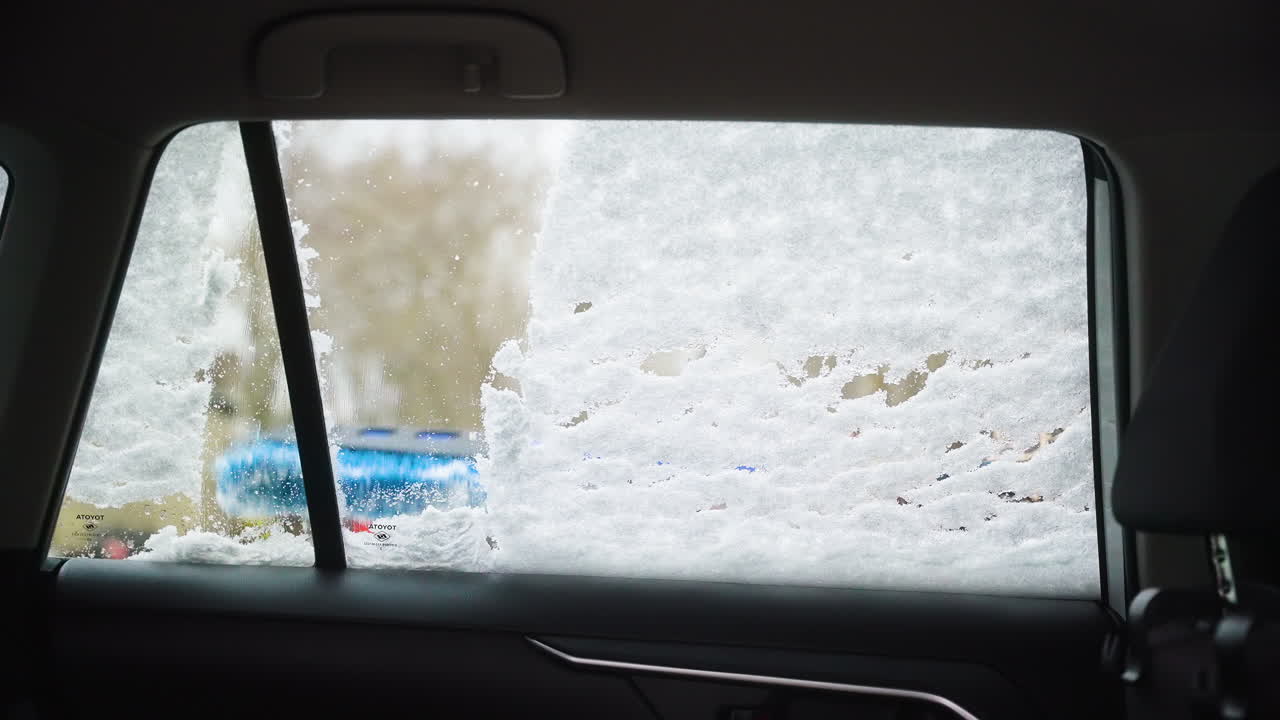 Close up of a car window covered in snow during cleaning, static shot showing brush wiping down the layer to reveal parking lot