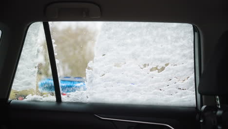 close up of a car window covered in snow during cleaning, static shot showing brush wiping down the layer to reveal parking lot
