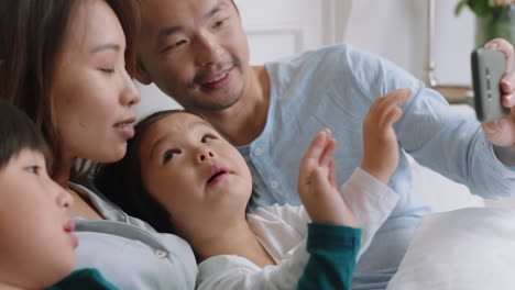 Feliz-Familia-Asiática-Teniendo-Video-Chat-Usando-Un-Teléfono-Inteligente-En-La-Cama,-Madre-Y-Padre-Con-Niños-Saludando,-Charlando-Con-Amigos-Por-Teléfono-Móvil,-Disfrutando-De-La-Comunicación-En-Línea,-Imágenes-De-4k