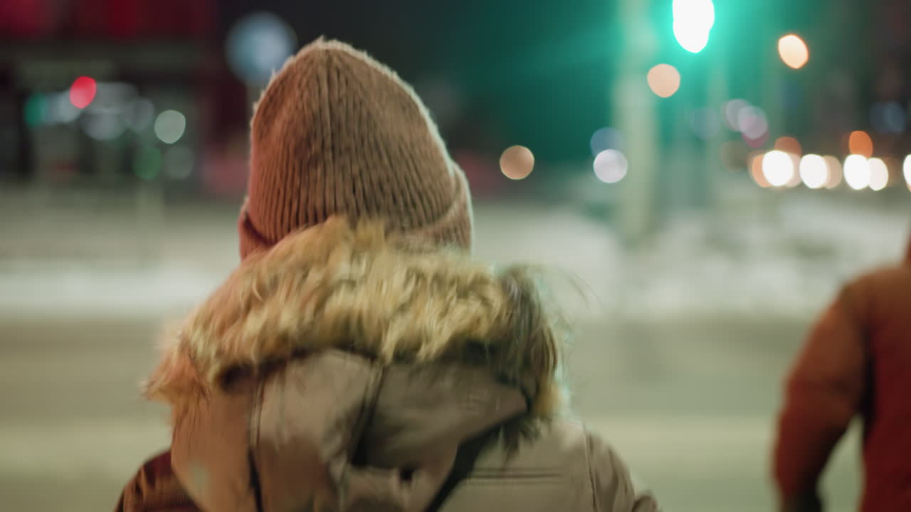 вид женщины в зимнем пальто с меховым капюшоном, стоящей перед зеленым светофором на ночном городском перекрестке с размытыми огнями и снежным фоном.