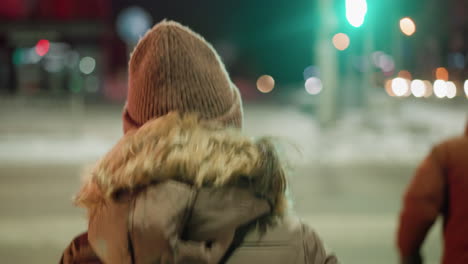 First-person view of a woman in a winter coat with a fur hood, facing a green traffic signal at a nighttime urban intersection with blurry lights and snowy background. Walking slowing across the road